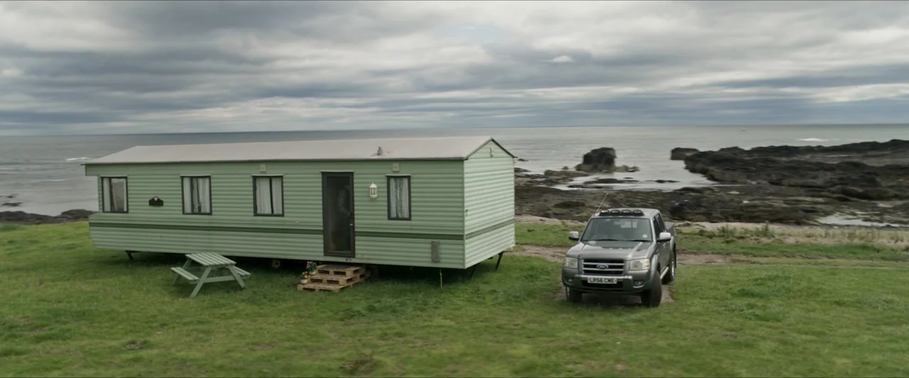 Last Breath (2025) movie still: wide — A light green mobile home sits on a grassy hill overlooking a rocky coastline. A gray pickup truck is parked…