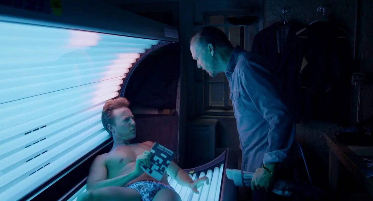Birdman (2014) movie still: medium — A dimly lit room contains a tanning bed with bright blue UV lights active. A man lies inside the tanning bed…