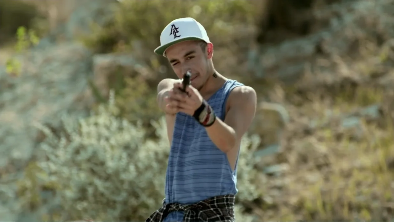 Loco Love (2003) movie still: medium — A dry hillside with sparse vegetation and rocky ground under bright daylight. A young man wearing a baseball…