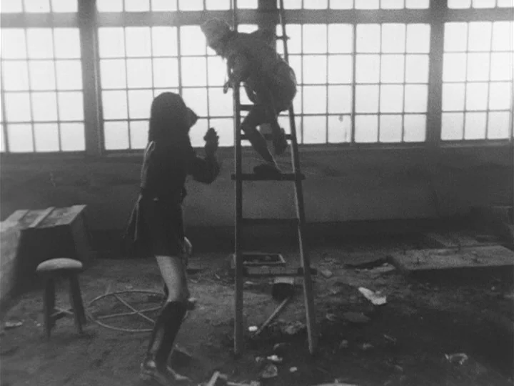 The War of Jan-Ken-Pon (1971) movie still: wide — A spacious industrial interior features grid windows and a cluttered dirt floor. One woman stands…