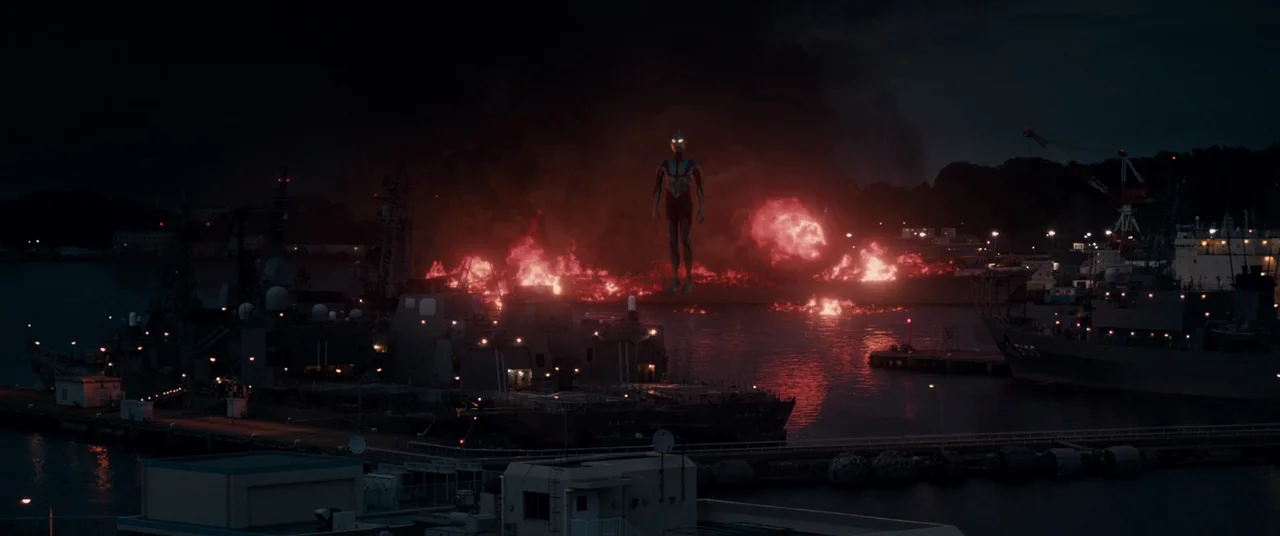 Shin Ultraman (2022) movie still: wide — A dark naval dockyard at night is engulfed in flames and thick black smoke. A singular humanoid giant…