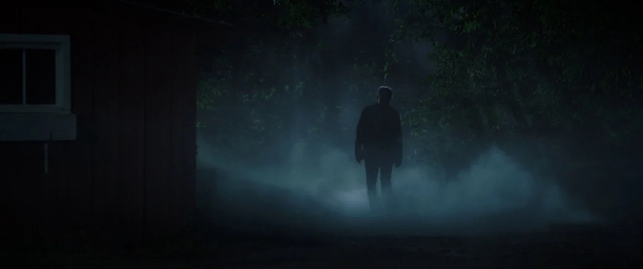 Pet Sematary (2019) movie still: wide — A dark, fog-filled wooded path beside the edge of a wooden cabin structure. A lone male figure walks forward…