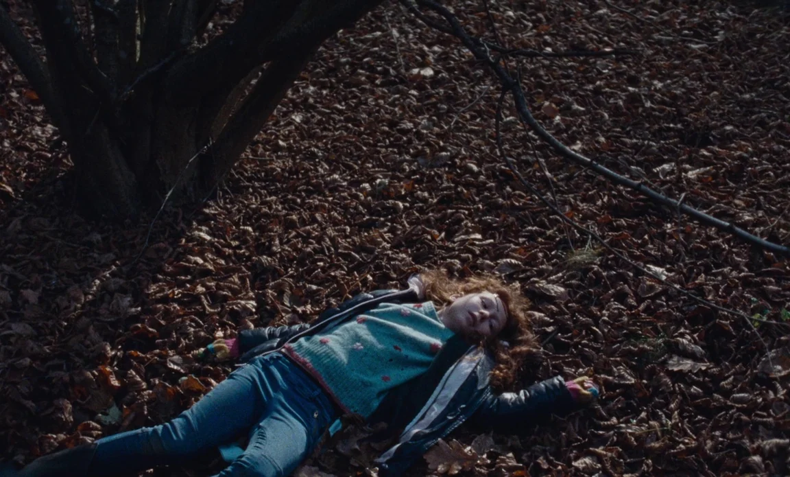High Life (2018) movie still: fullbody — A dense layer of fallen brown autumn leaves covers the woodland floor. A young girl lies flat on her back…