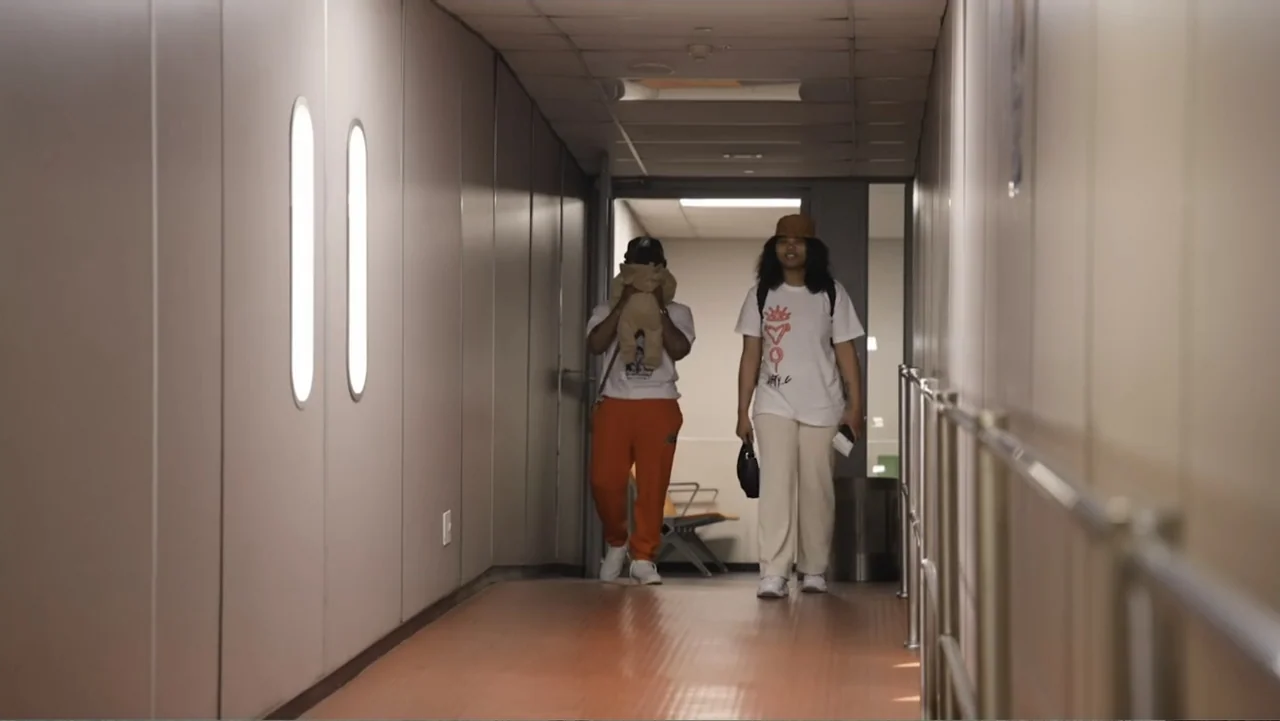 African Throne World Tour (2024) movie still: fullbody — A long interior pedestrian corridor with beige walls and orange flooring. Two young people…