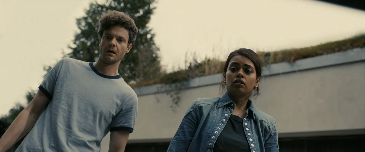 Companion (2025) movie still: medium — A man and a woman stand outdoors in front of a modernist structure. They look down with wide eyes at…