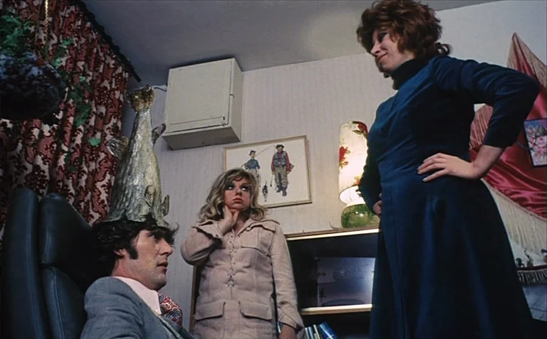 Business is Business (1971) movie still: medium — A living room interior featuring 1970s decor including floral curtains and framed wall art. A man…