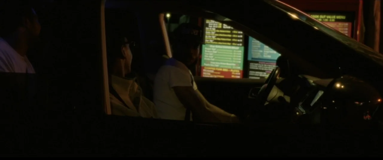 Sermon on the Mount (2019) movie still: medium — The interior of a car at night faces an illuminated drive-thru menu sign. Two men are seated inside…