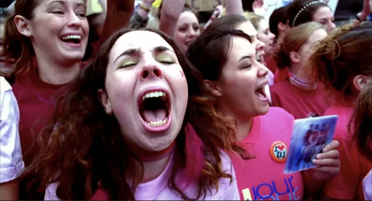 Josie and the Pussycats (2001) movie still: medium — A dense crowd of teenagers in matching pink t-shirts gathers at an outdoor event. Several young…