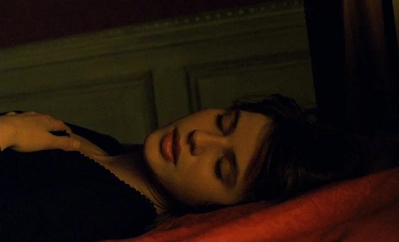 The Double Life of Véronique (1991) movie still: closeup — A woman lies on a bed with red sheets in a dimly lit room. She rests with her eyes…