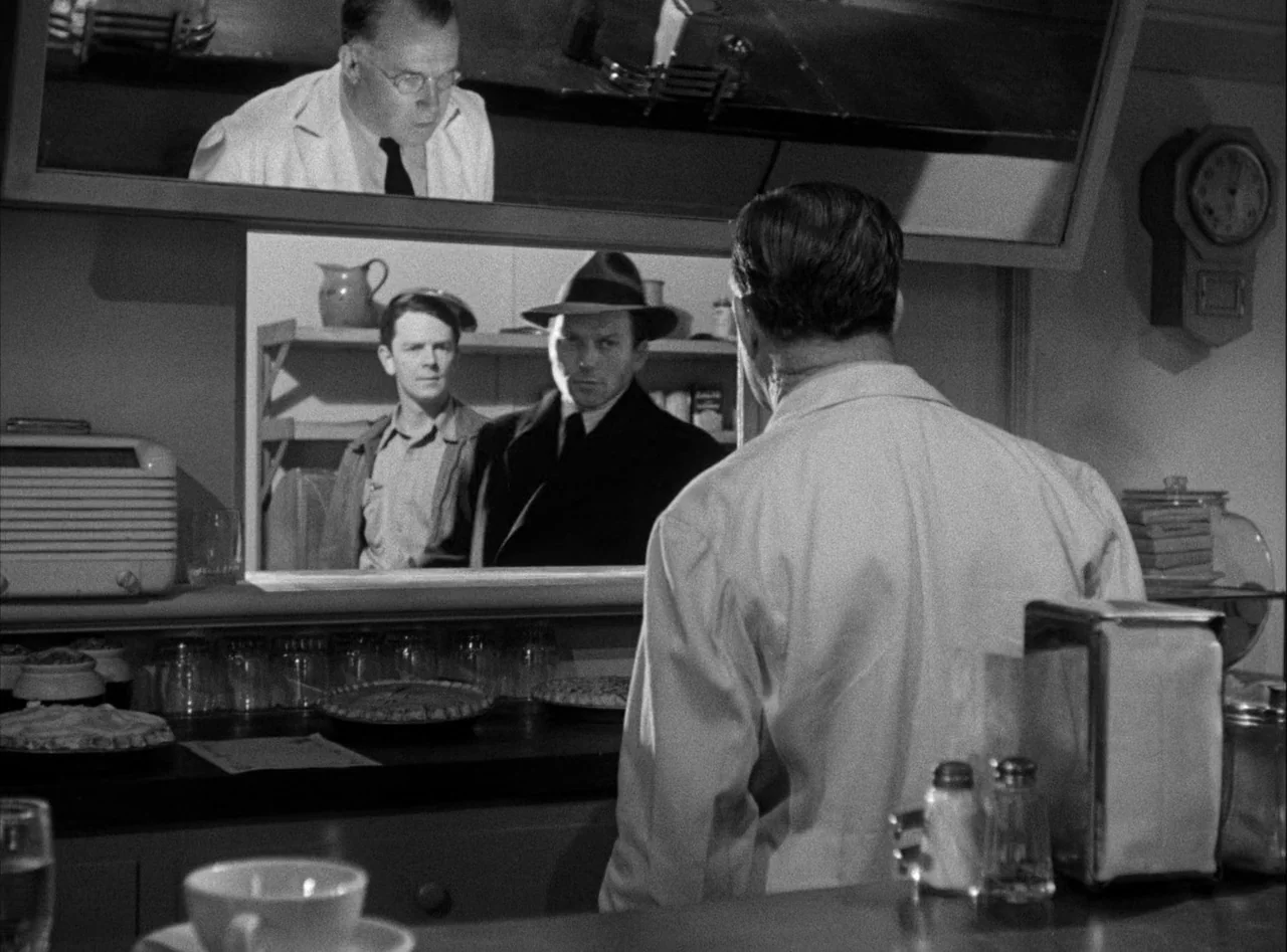 The Killers (1946) movie still: medium — A 1940s diner interior features a counter with pies and glasses in the foreground. A cook viewed from the…