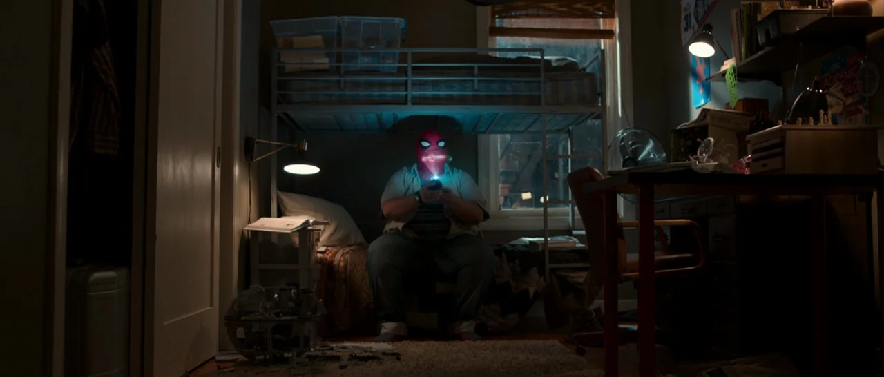 Spider Man: Homecoming (2017) movie still: medium — A dimly lit room contains a bunk bed and a cluttered desk. A person wearing a red mask sits on…