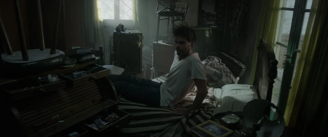 Terrified (2017) movie still: medium — A cluttered, dim space is packed with furniture and household items in disarray. A man sits upright on a bed,…