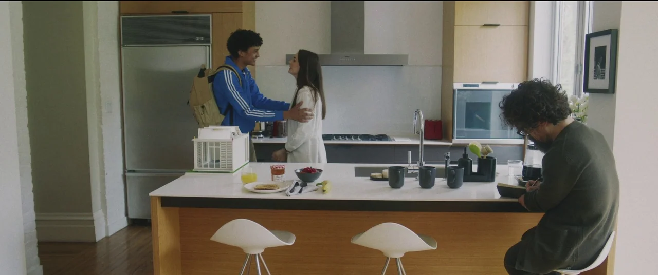 She Came To Me (2023) movie still: wide — A modern, bright home interior features light wood cabinetry and a central island workspace. Two young…