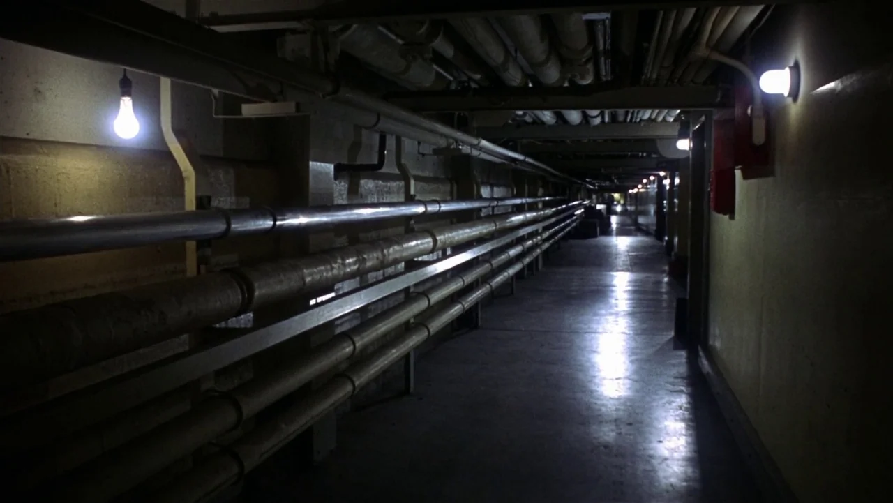 Bullitt (1968) movie still: establishing — An underground utility corridor features exposed piping running along walls and ceiling surfaces. The…
