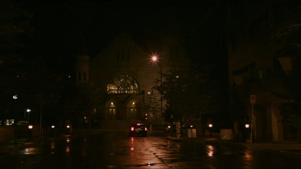 A Prairie Home Companion movie still: establishing — An empty wet asphalt street leads toward a large stone church building at night. A lone dark…