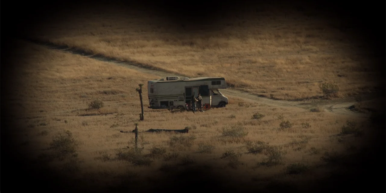 Fall (2022) movie still: wide — A vast, arid desert plain features dry brush and a worn vintage camper vehicle. Two individuals stand beside the…