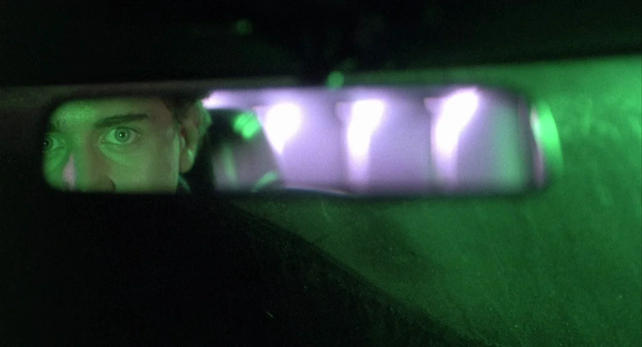 Vamp (1986) movie still: closeup — A close-up view through a horizontal rearview mirror inside a dark vehicle. A man's eyes and upper face are…