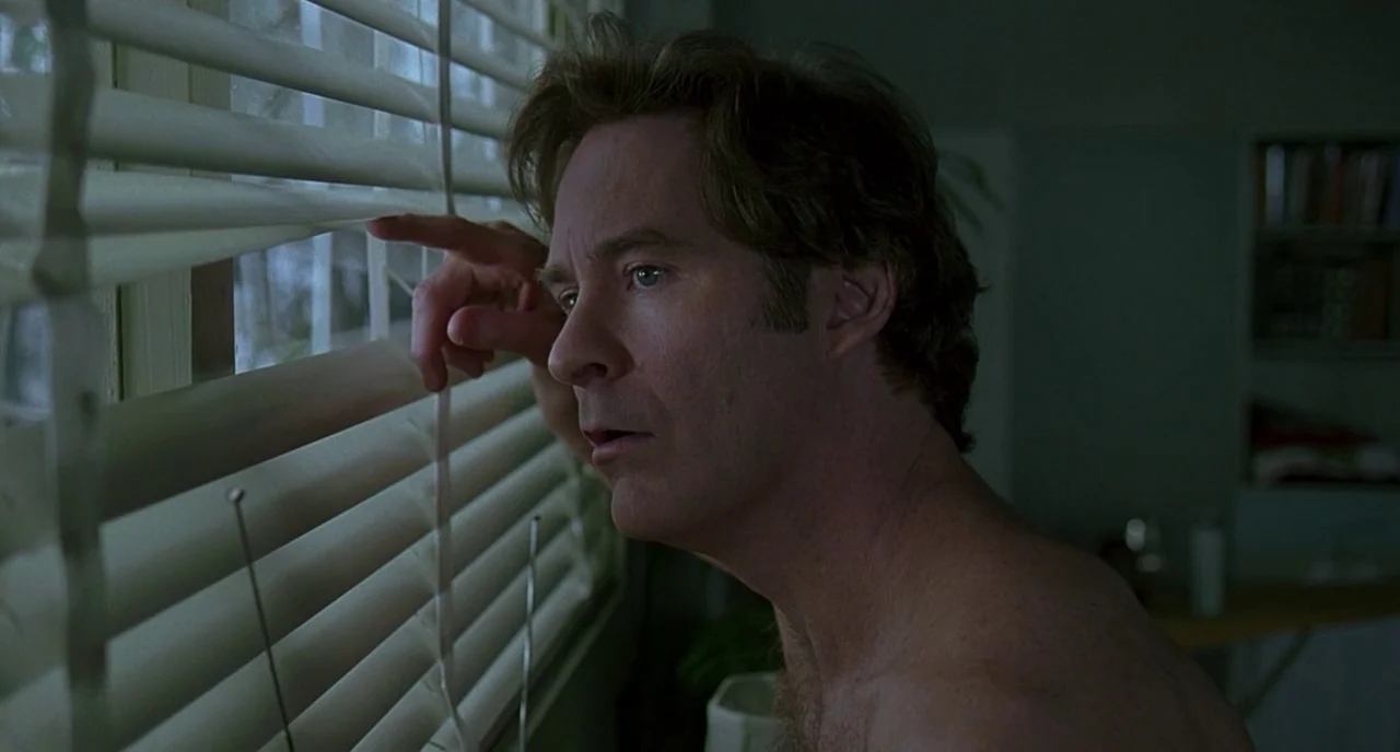 The Ice Storm (1997) movie still: closeup — A dimly lit room with bookshelves visible in the background and white window blinds. A shirtless man…