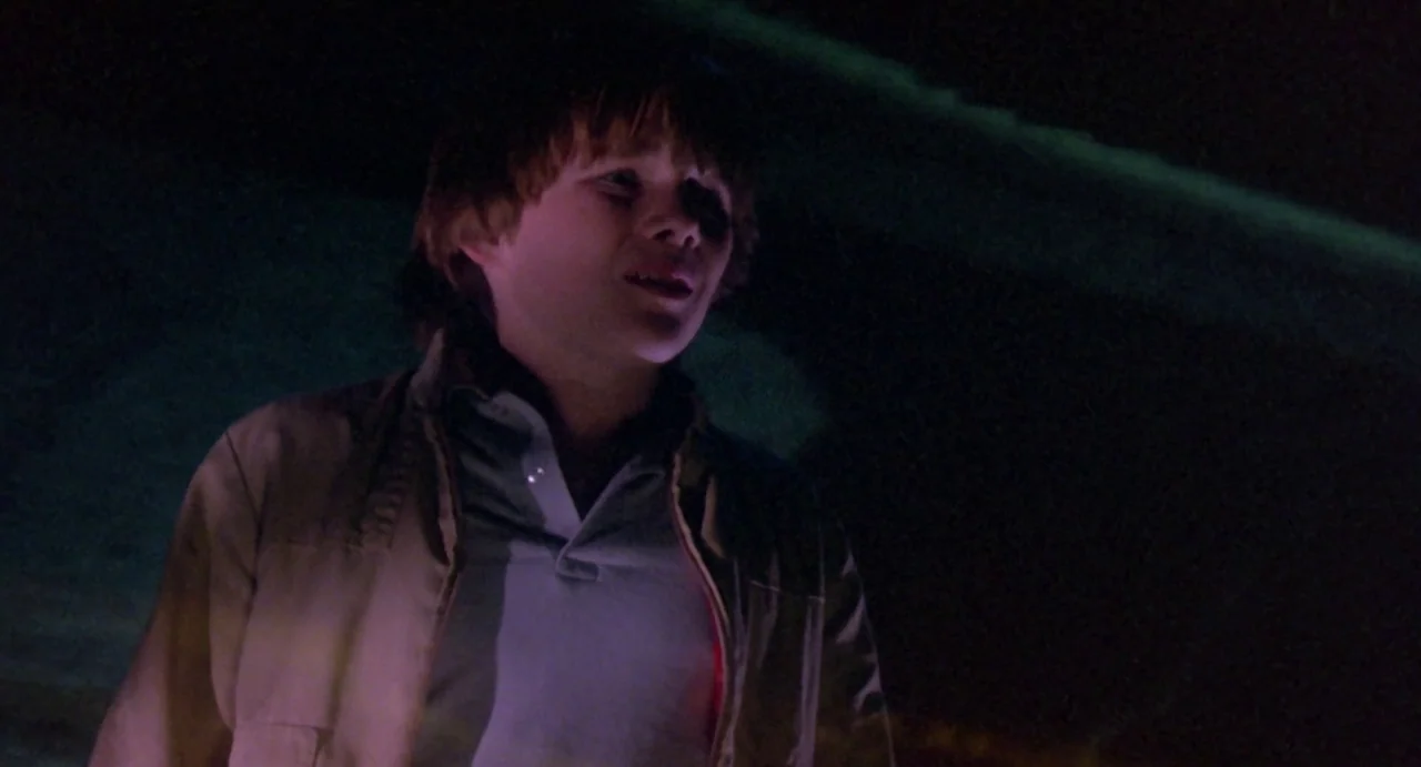 Explorers (1985) movie still: medium — A dark outdoor space illuminated by low-angle light reflecting from a nearby road surface. A young man stands…