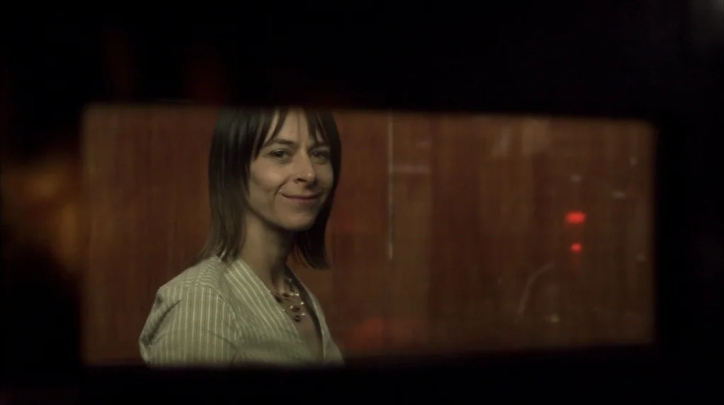 Red Road (2006) movie still: medium — A woman framed through a dark rectangular opening against a wood paneled wall. She stands in a room and looks…