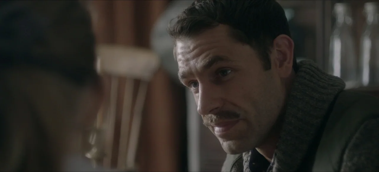 We Go In at Dawn (2020) movie still: closeup — A domestic interior with a person sitting at a wooden dining table. A man with a mustache looks…