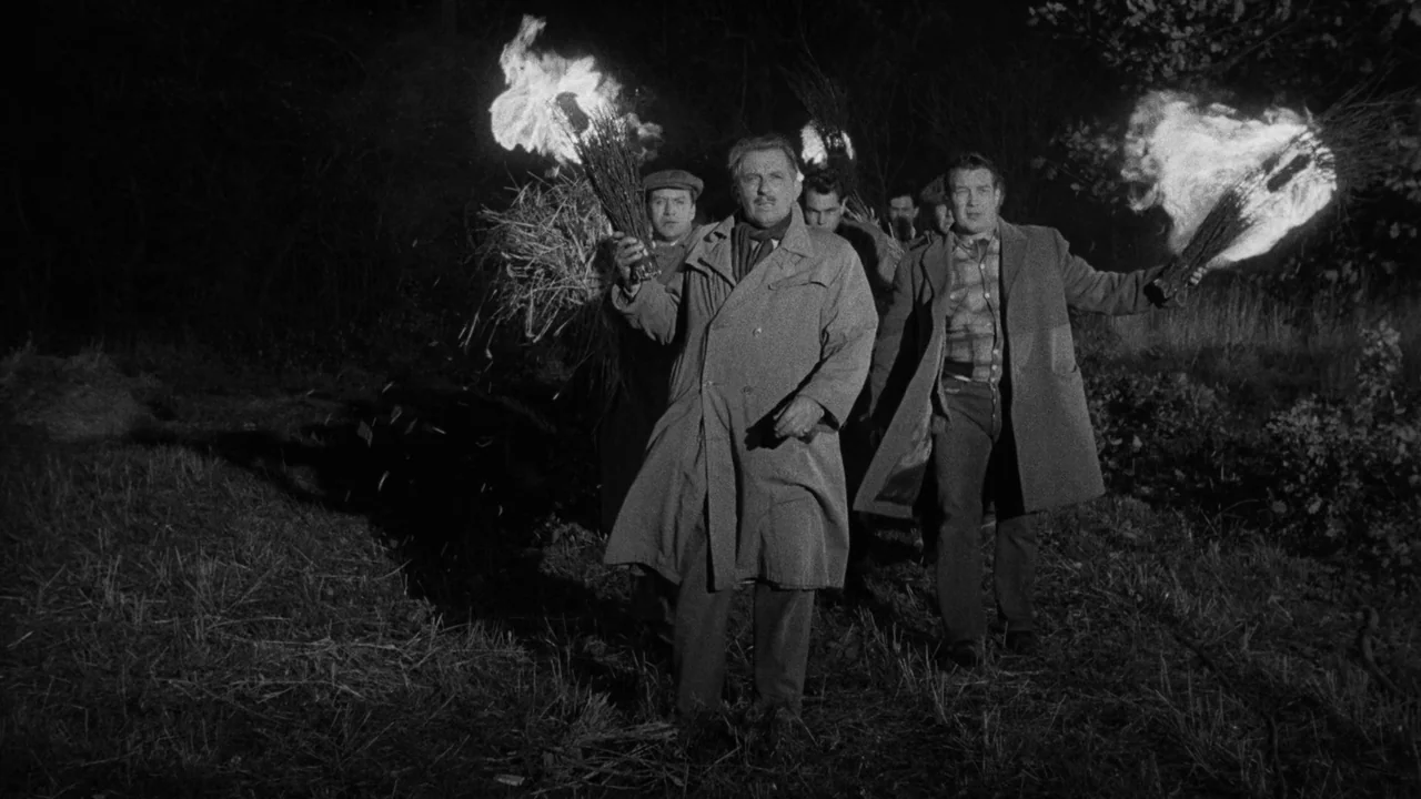 Village of the Damned (1960) movie still: fullbody — A dark, uneven field at night with scattered brush and tall grass. Several men walk forward…