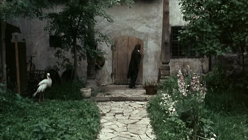 The Enigma Of Kaspar Hauser (1974) movie still: wide — A stone building with a wooden door sits behind a stone-paved path. A stork stands on the…