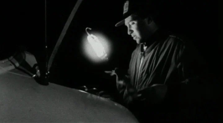 Roadkill (1989) movie still: medium — A dark outdoor space at night with the hood of a car raised open. A man wearing a cap looks down at the engine…