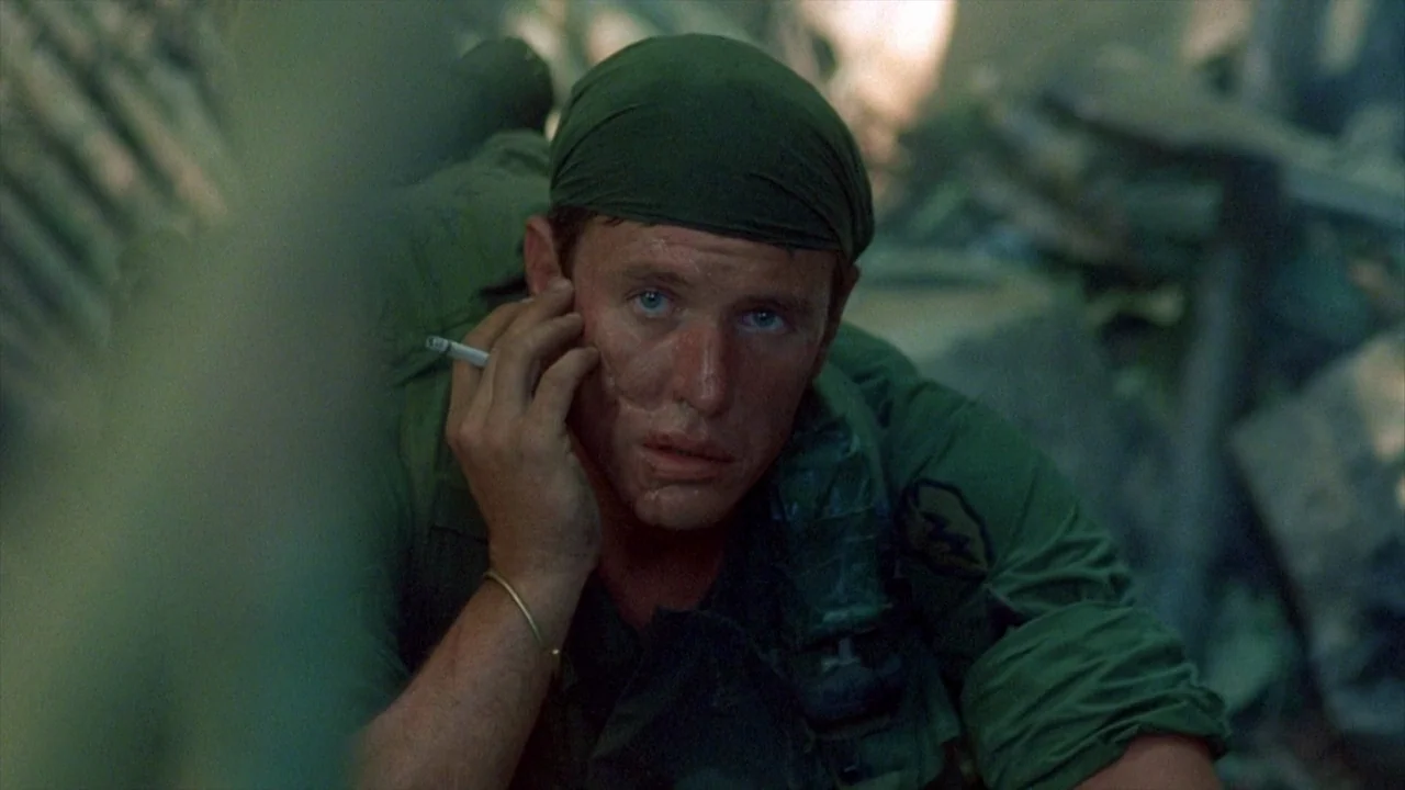 Platoon (1986) movie still: medium — Dense jungle foliage obscures the foreground and background of the outdoor setting. A young soldier in uniform…