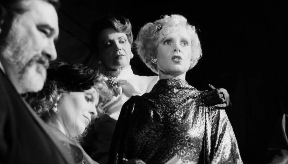 Veronika Voss (1982) movie still: medium — A dark, high-contrast interior space with multiple figures arranged in a cluster. The woman in the center…
