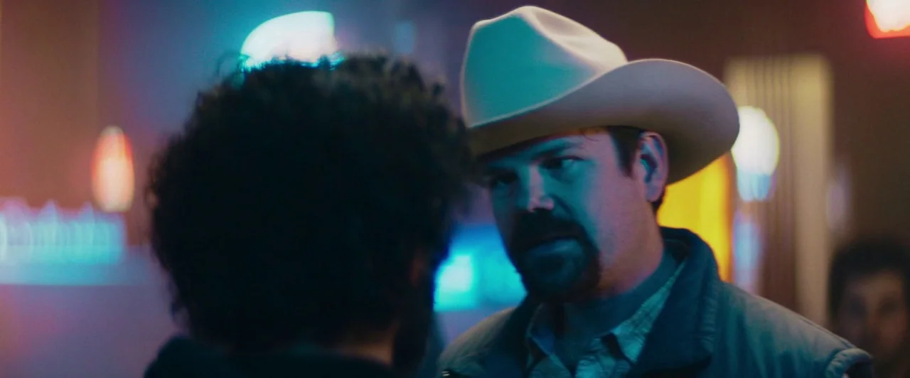 The Long Dumb Road (2018) movie still: medium — A dimly lit indoor space glows with colorful neon signage reflected on the surfaces. One man with a…