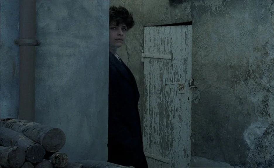 Au Revoir Les Enfants (1987) movie still: medium — A narrow stone alleyway features a weathered door with peeling paint and stacked logs. A young…
