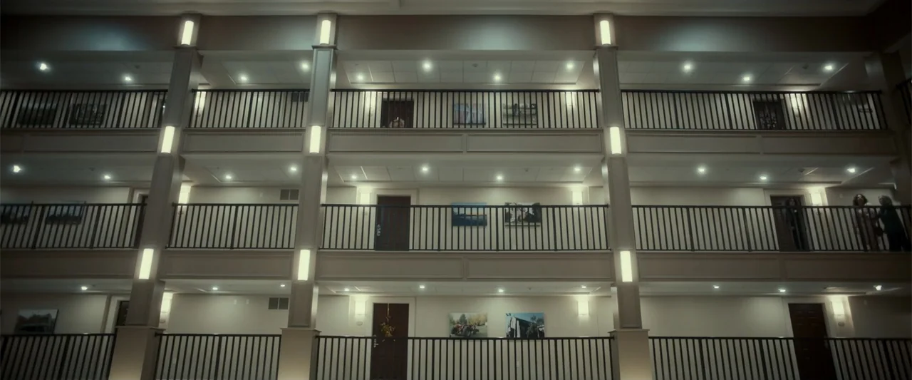 Jump, Darling (2022) movie still: wide — A multi-level hotel interior features stacked balcony walkways with metal railings and wooden doors. Art…