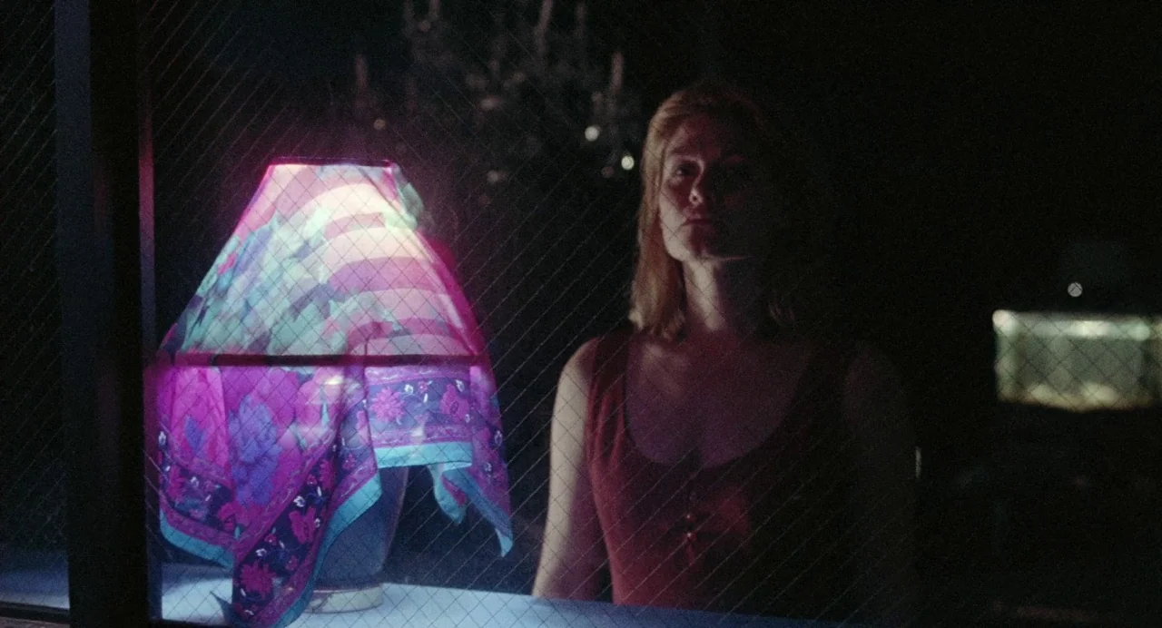What Happened Was… (1994) movie still: medium — A woman stands behind a wire-reinforced glass window in a dark room. Beside her, a lamp covered by a…
