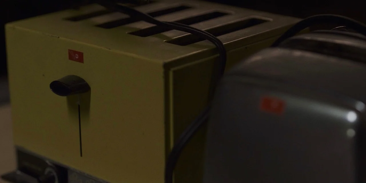 The Sound of Silence movie still: closeup — A close-up view shows a yellow box-shaped toaster beside a grey appliance. The toaster has a black lever…