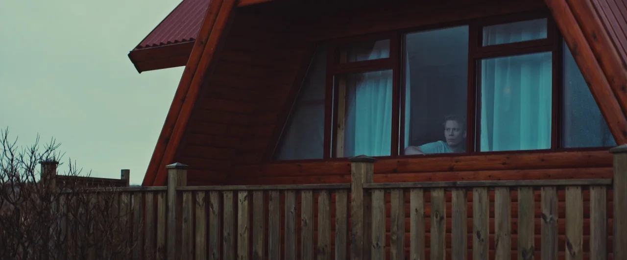 Rift (2017) movie still: wide — An a-frame wooden cabin stands against an overcast sky under a soft gray light. A young man looks out from the…