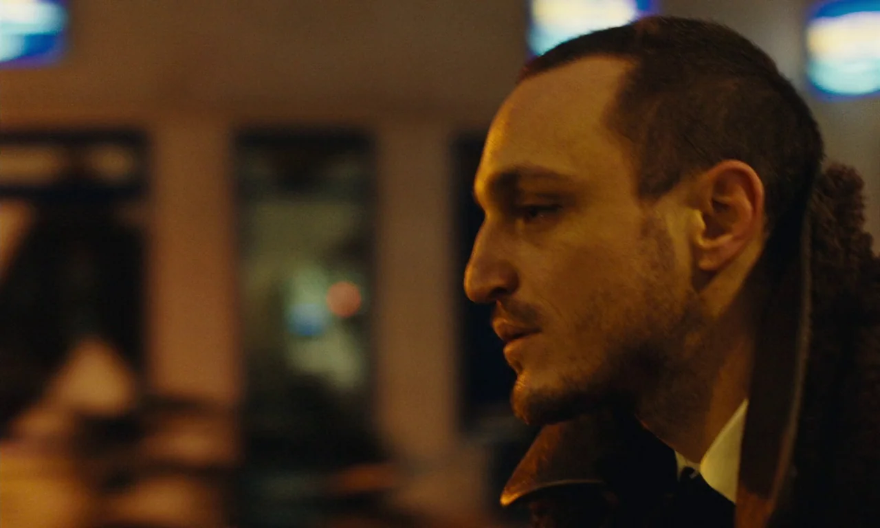 Passages (2023) movie still: closeup — A dimly lit indoor space featuring blurred, glowing blue lights in the background. A man stands in profile…