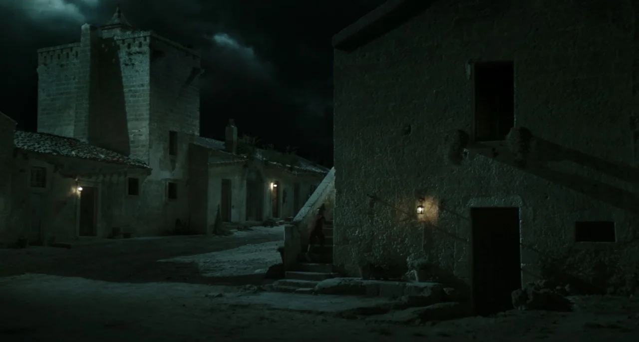 Pinocchio (2022) movie still: wide — A dark medieval village square is composed of rough stone buildings under a cloudy night sky. A small child…
