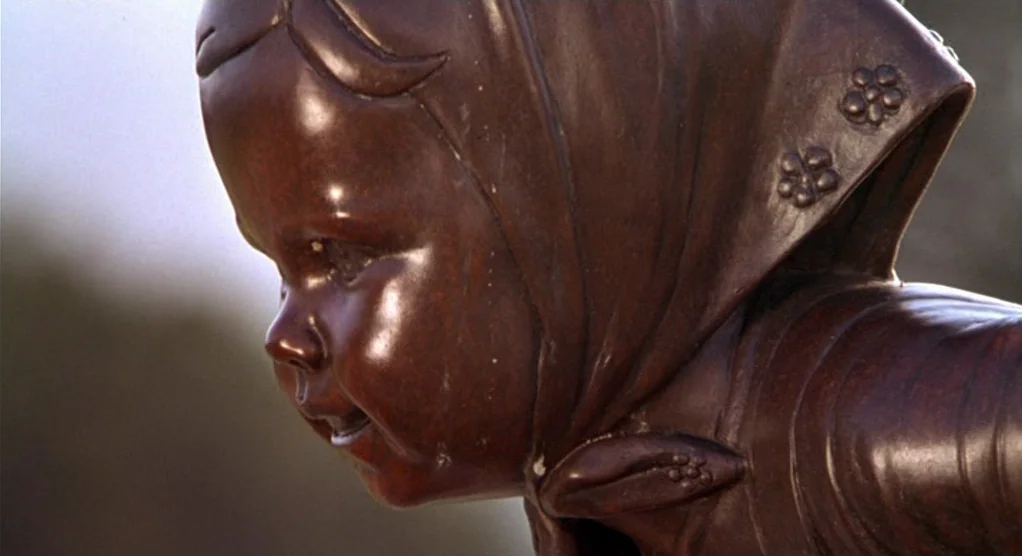 The Crossing Guard (1995) movie still: closeup — A close-up view shows a bronze sculpture of a child wearing a bonnet. The statue stands fixed…