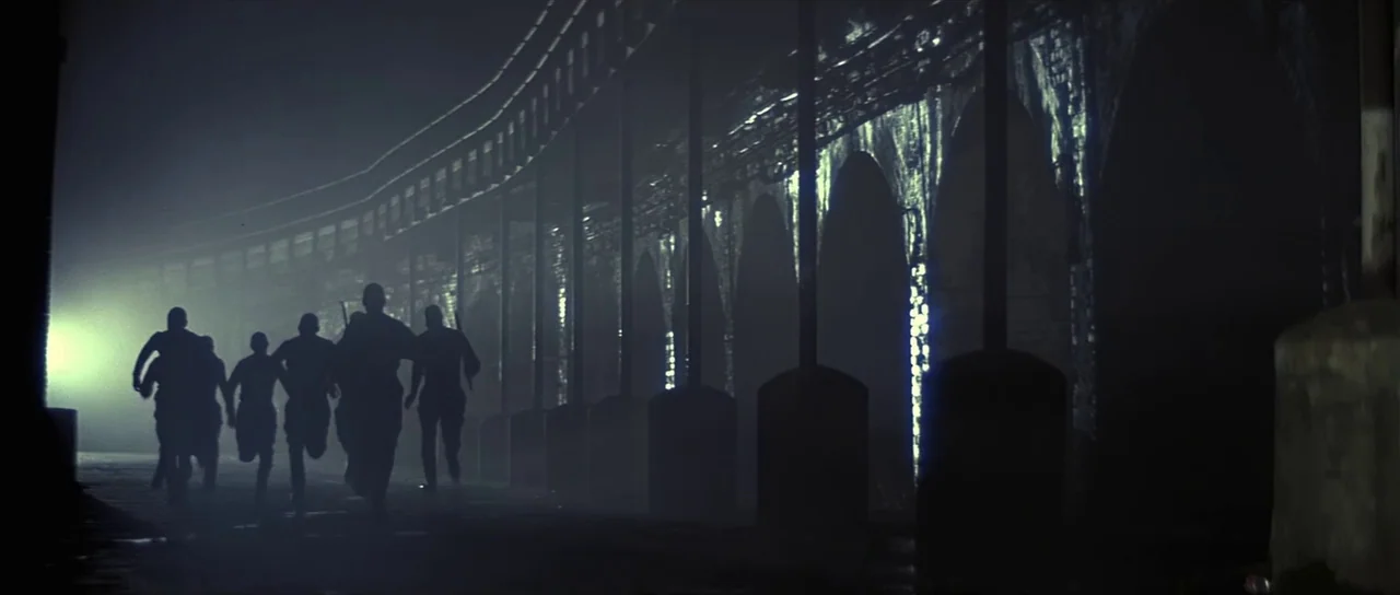 Pink Floyd The Wall (1982) movie still: wide — A dark brick viaduct at night with repeated arches and glowing streetlamp light. Five men run away…