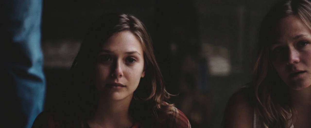 Martha Marcy May Marlene (2011) movie still: medium — An interior space with dim, low-contrast lighting frames two young women in conversation. The…