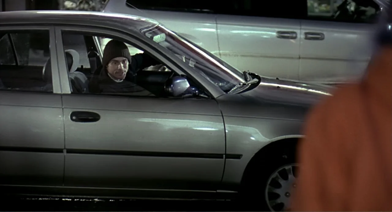 Eternal Sunshine of the Spotless Mind (2004) movie still: medium — A silver sedan parked in a dimly lit outdoor lot at night. A man sits in the…