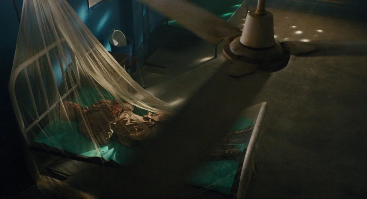 Submergence movie still: medium — A dimly lit room contains a single hospital bed draped with a sheer white net. An elderly man lies in the bed with…