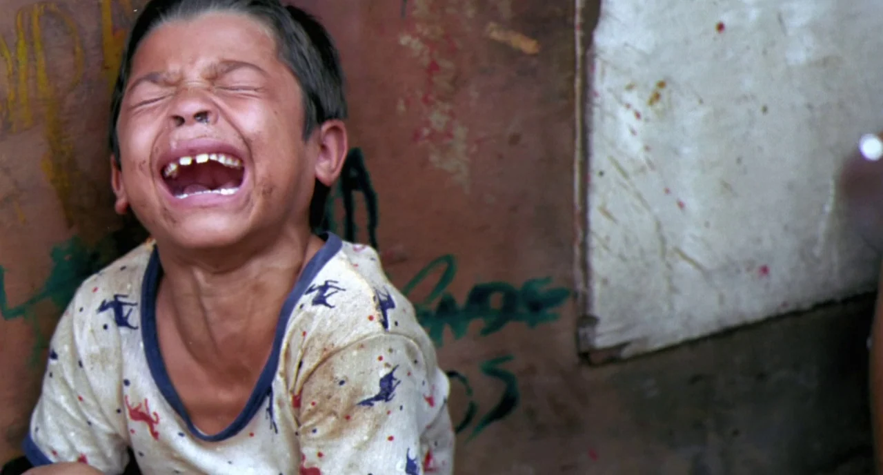 City of God (2002) movie still: closeup — A boy stands in front of a textured wall covered in dark graffiti markings. The boy yells loudly with his…
