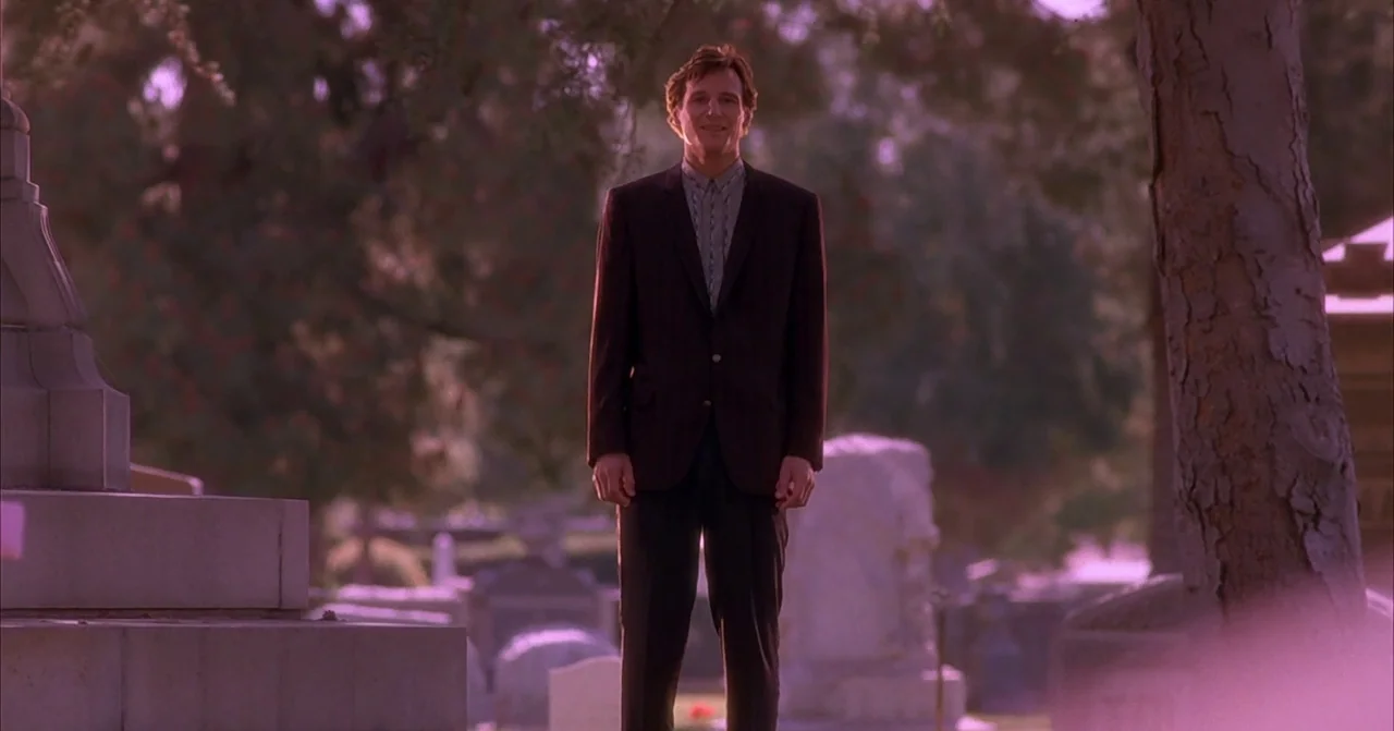 Darkman (1990) movie still: fullbody — A cemetery featuring numerous stone grave markers and mature trees at golden hour. A man in a dark blazer…