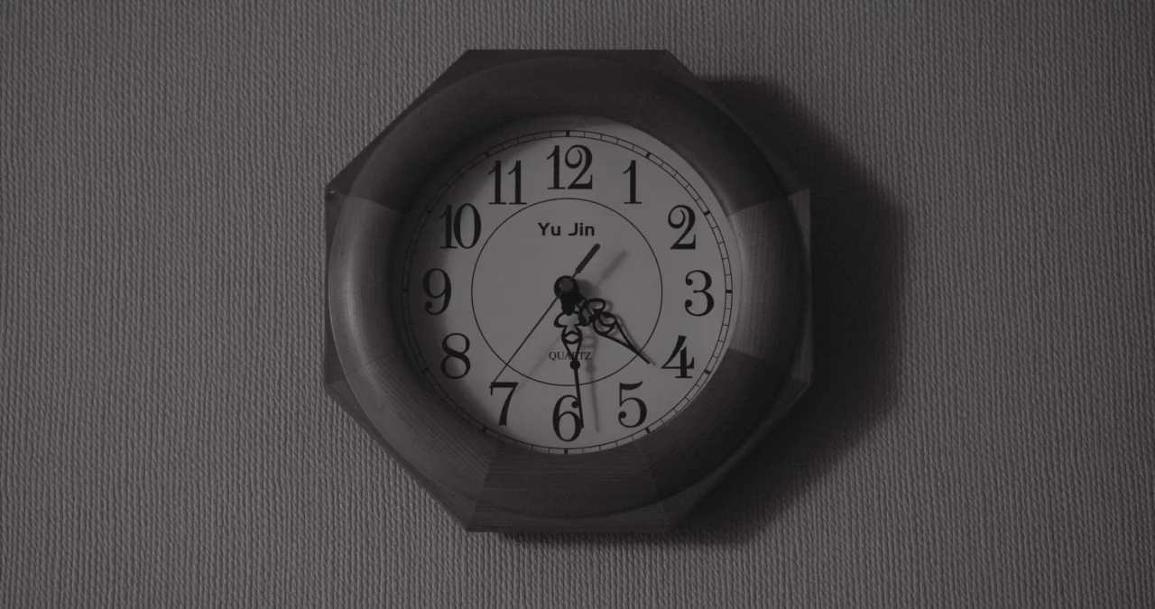 The Day After movie still: closeup — An octagonal analog clock with a wooden frame hangs on a textured wall. The clock face displays the time as…