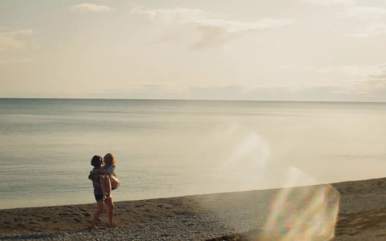 Knocking (2021) movie still: wide — A vast, calm ocean stretches toward a low horizon under a hazy sky. A person carries another person in their…