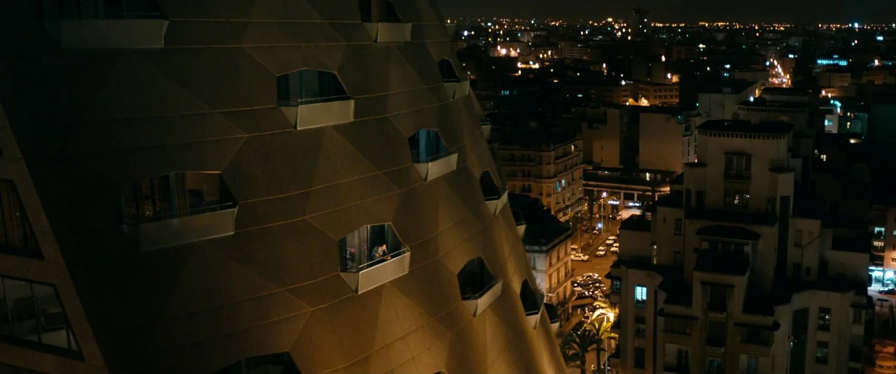 A Hologram for the King movie still: wide — A high-angle view of a modern skyscraper with faceted brown cladding at night. A lone figure stands on…