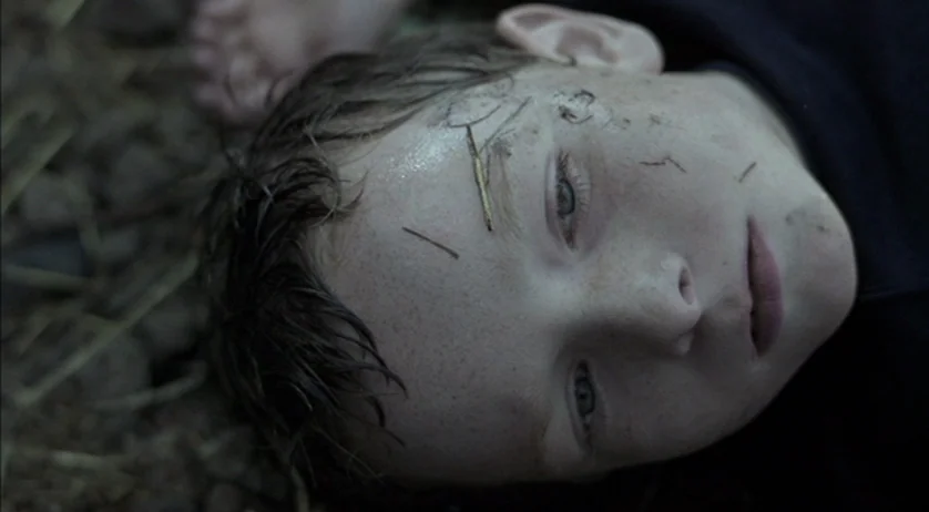Ratcatcher (1999) movie still: closeup — A child lies flat on dark, damp soil with scattered dry pine needles. The boy rests on the ground while…