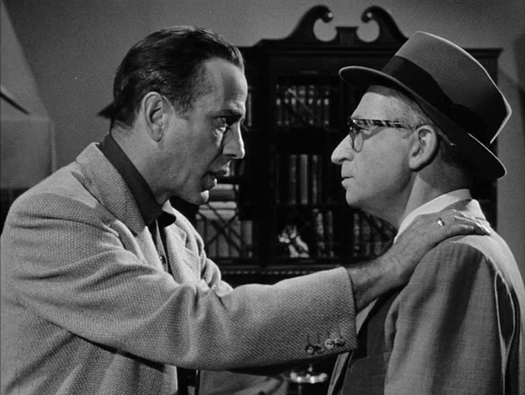 In a Lonely Place (1950) movie still: medium — A dark study contains a large wooden cabinet filled with books in the background. A man grips the…